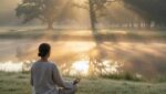 A person meditating cross-legged outdoors at sunrise, surrounded by soft light and gentle mist, representing calmness and mindfulness.